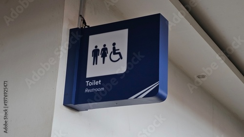 Valokuva Blue restroom sign mounted on wall showing male, female, and wheelchair icons, with “Toilet Restroom” text, hanging bracket, and ceiling structure visible