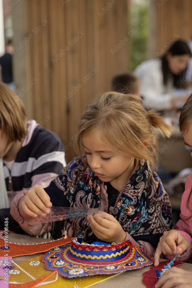 Fototapeta premium Girl creates craft with colorful patterns outdoors Description,Young girl concentrates on crafting activity, arranging colorful elements on decorative piece.