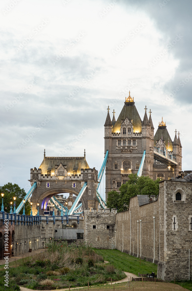 Fototapeta premium The northern Victorian Gothic tower and its adjacent arch stand bathed in warm, golden light, creating a sharp contrast against the cool, overcast sky. Vibrant blue suspension cables offer a striking 