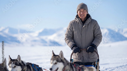 Man in gray winter coat stands on snow-covered landscape with sled dogs, mountains visible in the background under clear blue sky