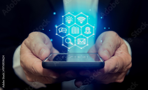 Businessman holding a smartphone with glowing digital business icons hologram, representing technology, data, communication, and modern networking on dark background.