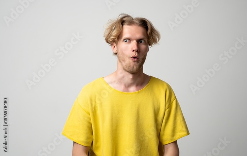 Canvas Print Young blond man in a yellow t-shirt expressing shock and surprise, widening eyes