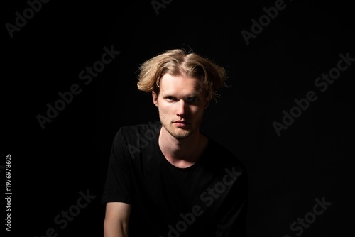 Young man in a black t-shirt facing the camera on a dark background with dramatic lighting highlighting his serious expression, conveying mystery and mood