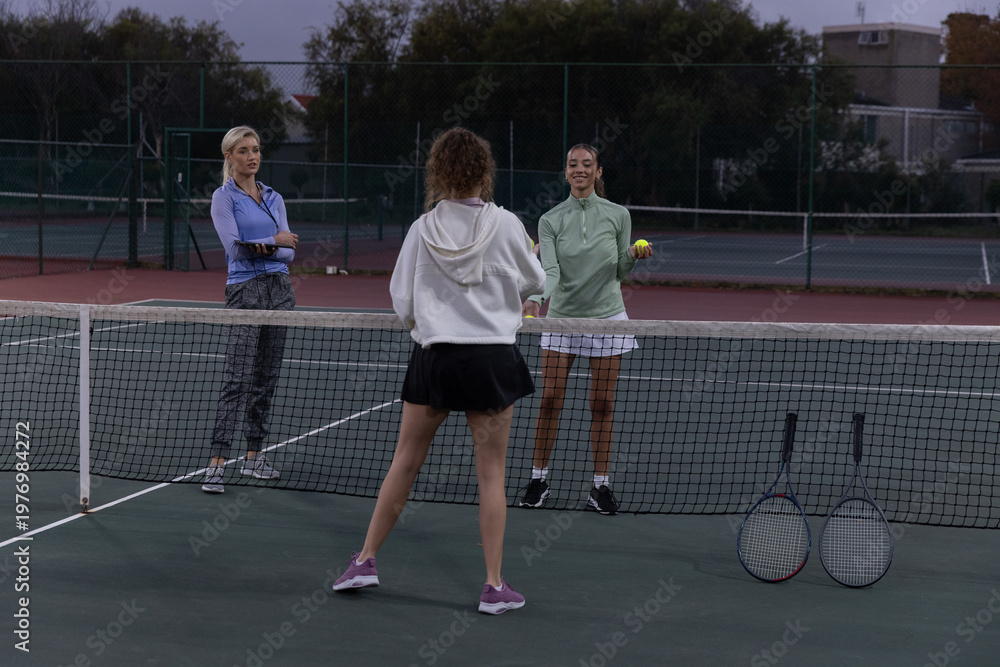 Obraz premium Female players practicing tennis at dusk on court, wearing hoodies, holding tennis ball and rackets