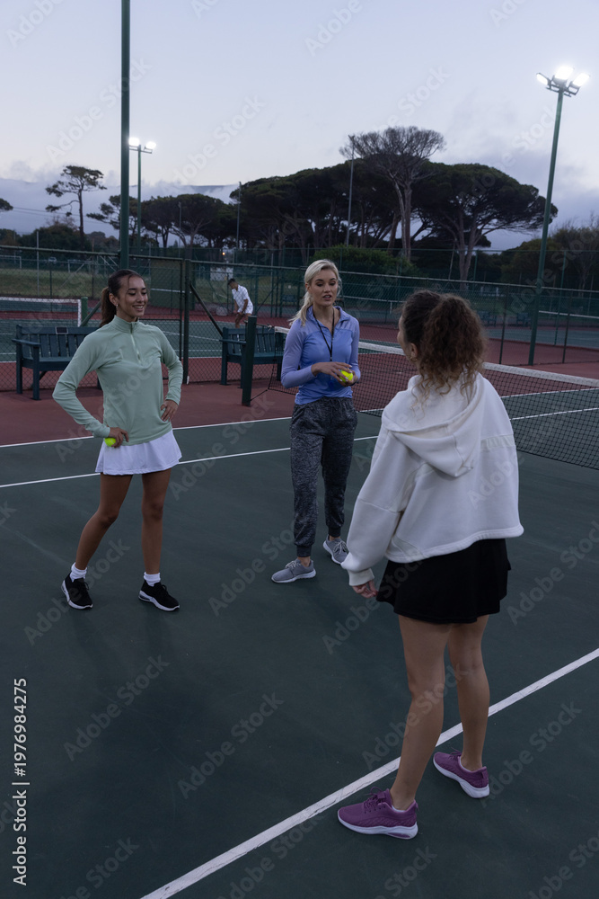 Obraz premium Female friends standing on fenced court at dusk holding yellow tennis balls near net in skirts