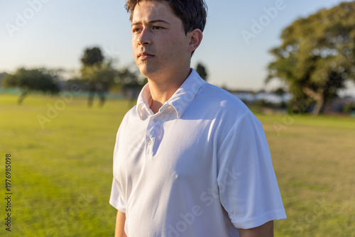 Man standing and looking right across grassy park during golden hour, wearing white polo shirt
