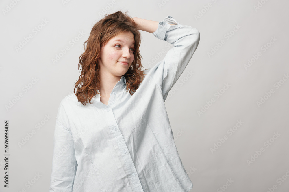 Naklejka premium Young woman with curly hair wearing a white shirt looks to the side with a slight smile, hand resting on her head. Neutral background, casual lifestyle and natural expression concept.