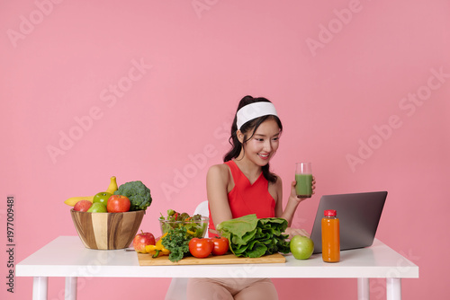 Healthy nutrition and digital lifestyle concept with Asian woman drinking green smoothie at table with fresh vegetables and laptop diet planning wellness routine and clean eating habits