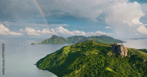 Yasawa Islands, Fiji: striking colorful rainbow arching over lush green volcanic mountains and pristine Pacific Ocean, revealing a tropical paradise nature landscape. Aerial view drone flight panorama
