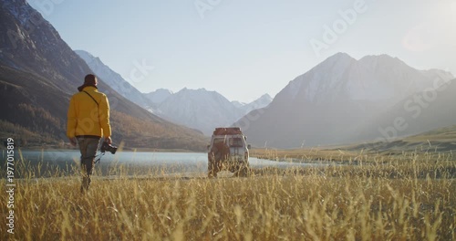 Traveler in a yellow jacket carrying a camera and walking toward an off road SUV parked by a mountain lake in a remote alpine valley. Golden grass, snowy peaks, and bright sunlight create a scenic