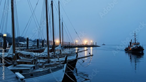 Early Morning Harbor Scene with Sailing Vessels and Tugboat Under Hazy Blue Skies and Pier Lights