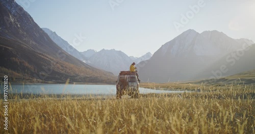 Traveler in a yellow jacket relaxing on the roof of a parked off road car and making a smartphone photo by a mountain lake. Snow covered peaks, golden meadow, and bright natural light create a