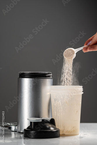 close-up shot of a woman's hand using a plastic scoop to pour whey protein powder into a transparent shaker bottle. A silver supplement container and a black lid are on a white table against a grey ba