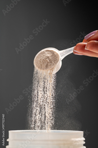 close-up shot of a woman's hand using a plastic scoop to pour whey protein powder into a transparent shaker bottle. A silver supplement container and a black lid are on a white table against a grey ba