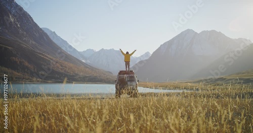 Traveler in a yellow jacket standing on the roof of an off road SUV with raised arms near a mountain lake in a remote alpine valley. Snowy peaks, golden grass, and bright sunlight create a powerful