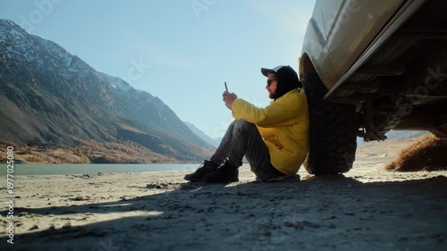 Male traveler in a yellow jacket using a smartphone while sitting on the ground and leaning against an off road SUV near a mountain lake. Scenic outdoor setting with alpine slopes, open shoreline, and