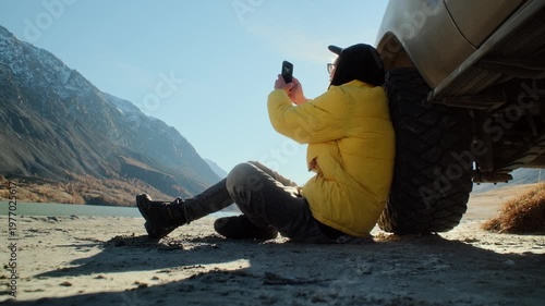 Male traveler in a yellow jacket taking a smartphone photo while sitting on the ground and leaning against an off road SUV near a mountain lake. Scenic outdoor setting with alpine slopes, open