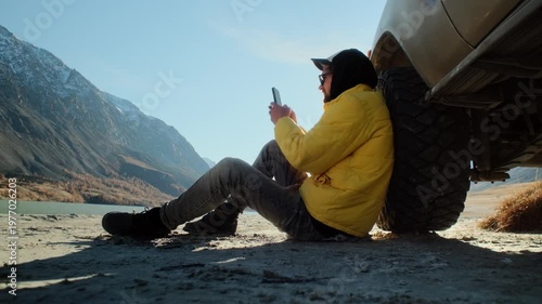 Traveler in a yellow jacket making a phone photo while sitting by a parked SUV on the shore of a mountain lake. Calm water, alpine terrain, and bright sunlight create a strong commercial scene about