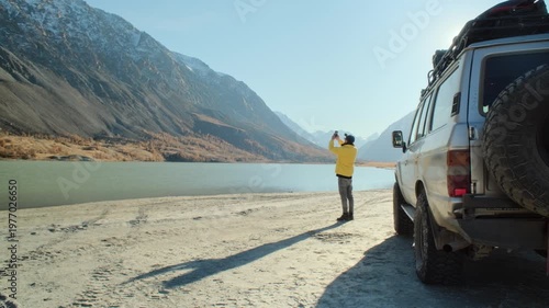 Male traveler in a yellow jacket taking a smartphone photo beside an off road SUV parked near a mountain lake. Snowy slopes, open shoreline, and bright daylight create a commercial outdoor travel