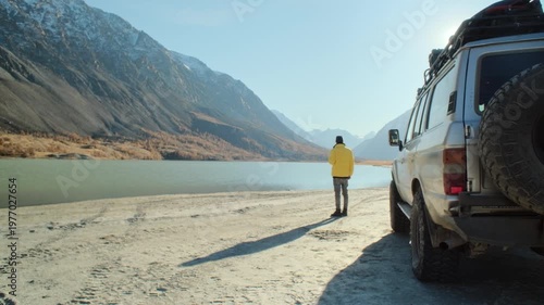 Man in a yellow jacket holding a coffee mug near an off road vehicle on the shore of a mountain lake. Wide alpine landscape, bright sun, and remote nature create a strong commercial image about