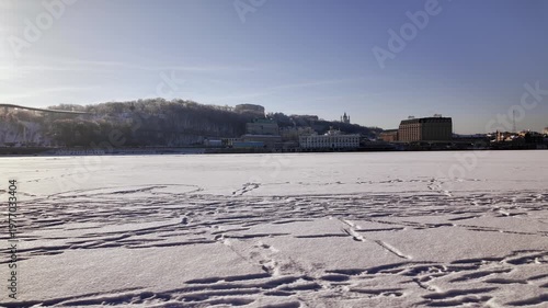 Static wide landscape of frozen Dnieper river covered in snow in Kyiv. View of Podil embankment with city buildings and smoking chimneys under clear blue sky.