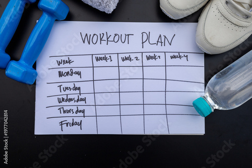 Top view of a blank weekly workout plan sheet on black floor with dumbbells, sneakers, and water bottle