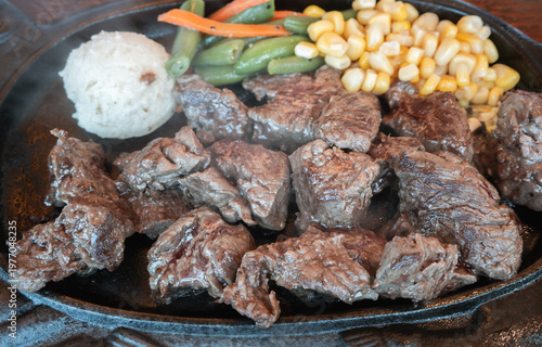 Sizzling diced beef steak with vegetables on black iron plate.