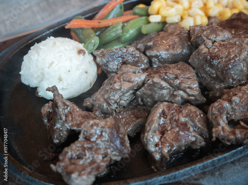Sizzling diced beef steak with vegetables on black iron plate.