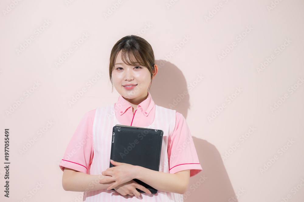 Fototapeta premium ピンク背景で介護士や保育士として働く女性の訪問介護とやりがいある仕事のイメージ 転職などのイメージにカメラ目線の笑顔 タブレットを持つ