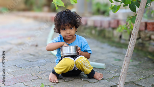 Fotografie Portrait of a poor Indian child sitting barefoot on the pavement, holding and eating from a metal bowl