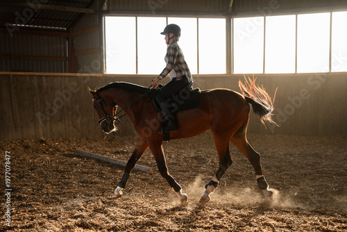 Female rider training bay horse in indoor arena at sunset. Equestrian sport and dressage concept, professional horse riding school, athlete practicing , harmony and discipline, equestrian training