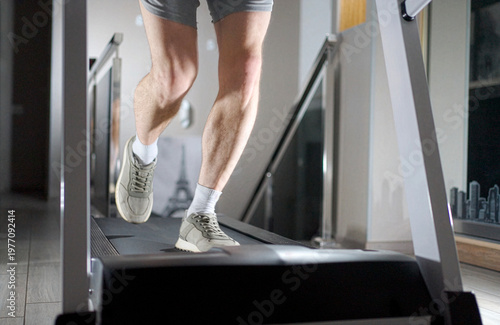 Close-up of Man Legs Running on Treadmill at Home. A man in sneakers and shorts running on a treadmill. Cinematic low angle shot focusing on leg muscles and movement during a cardio workout