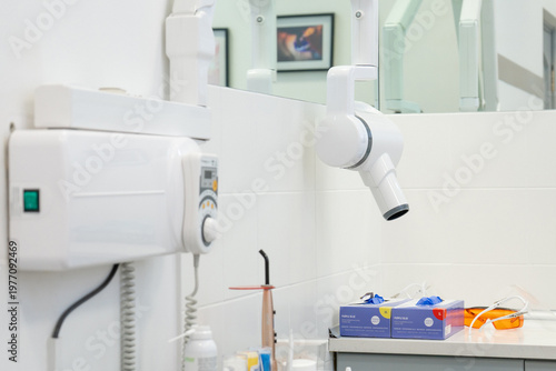 Wall mounted dental x-ray equipment in bright medical office. Professional diagnostic tool for teeth radiography with protective gloves and glasses on background.