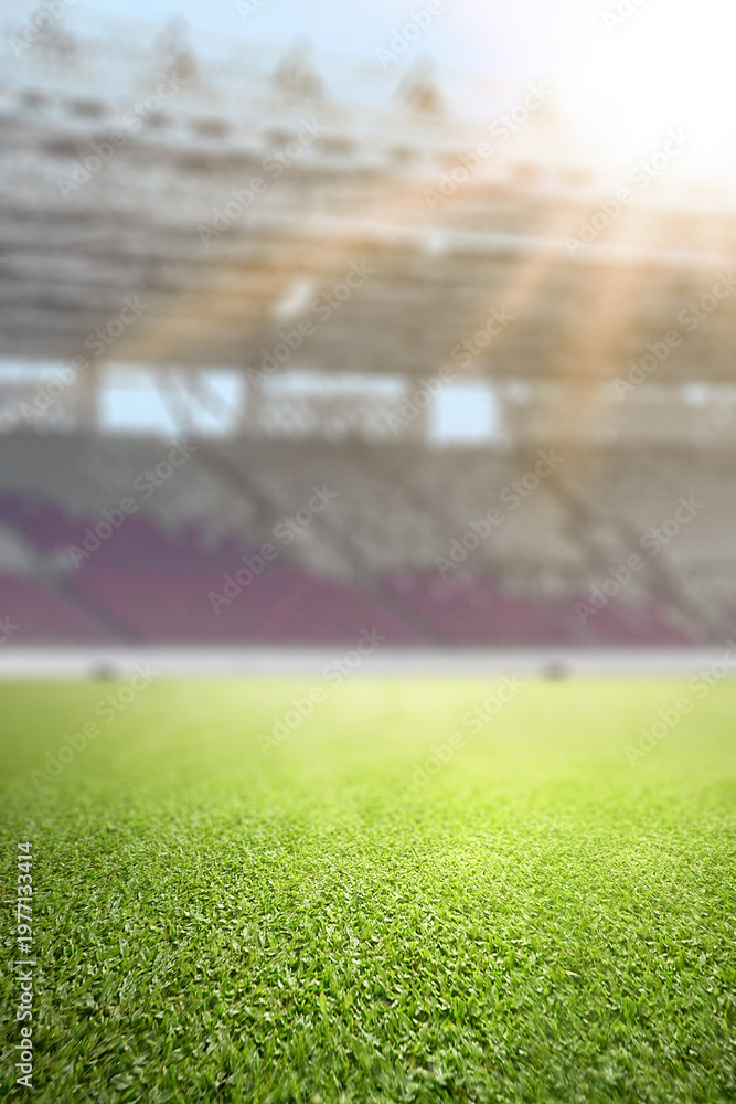 Fototapeta premium Close-up of lush green grass on a professional soccer field with a blurred stadium and bright sunlight in the background. Ideal for sport-themed designs