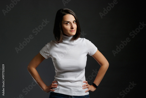 A neutral portrait of a young woman with a steady gaze standing in a white turtleneck against a dark background.