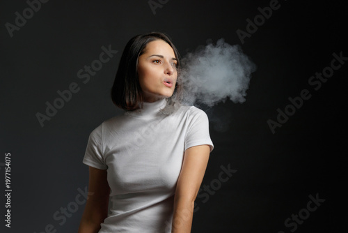 Portrait of a woman in a white turtleneck exhaling vapor from a vape pen, looking to the side against a dark studio background.