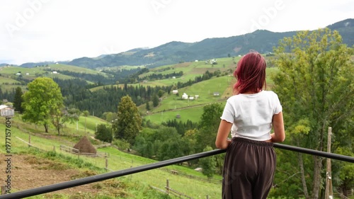 Girl stands on a balcony and looks over the mountains and fields in a rural area in summer