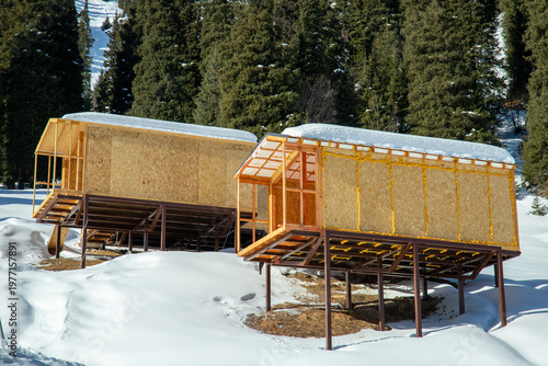 Two modular buildings made of OSB with visible yellow mounting foam insulation on a metal frame base in a winter mountain landscape.