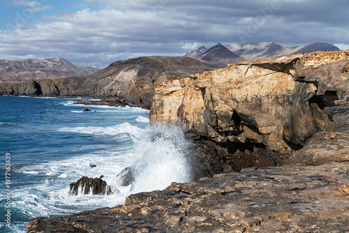 Powerful waves crash against rugged coastal cliffs in Jandia, creating a dramatic seascape with dark rocks, distant mountains, and moody clouds above the Atlantic, Fuerteventura, Canary islands, Spain