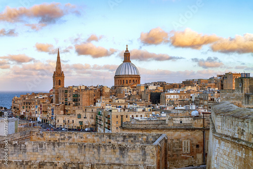 Elevated view of a historic coastal city with Cathedral of Saint Paul, warm stone buildings, and soft clouds glowing in gentle evening light, Valletta, Malta.