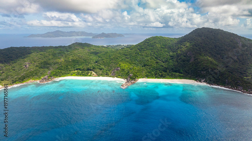 Turquoise waters meeting tropical green hills on a sunny coastline. La Digue, Seychelles.