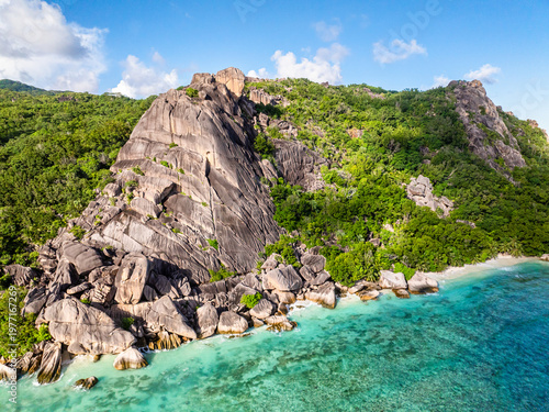 Imposing rocky cliffs rise above lush greenery, with turquoise waters at the base. Seychelles, La Digue.