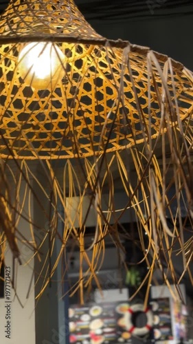 Handmade wicker straw pendant lamp glowing in a tropical restaurant at night in Phuket, Thailand. Exotic boho style light fixture with long fringes in a cozy evening cafe atmosphere during vacation.