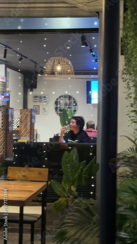 Cozy evening cafe interior with a stylish wicker straw lamp, green tropical plants and blurred city lights through the window. Warm atmosphere of a modern restaurant at night with soft lighting.