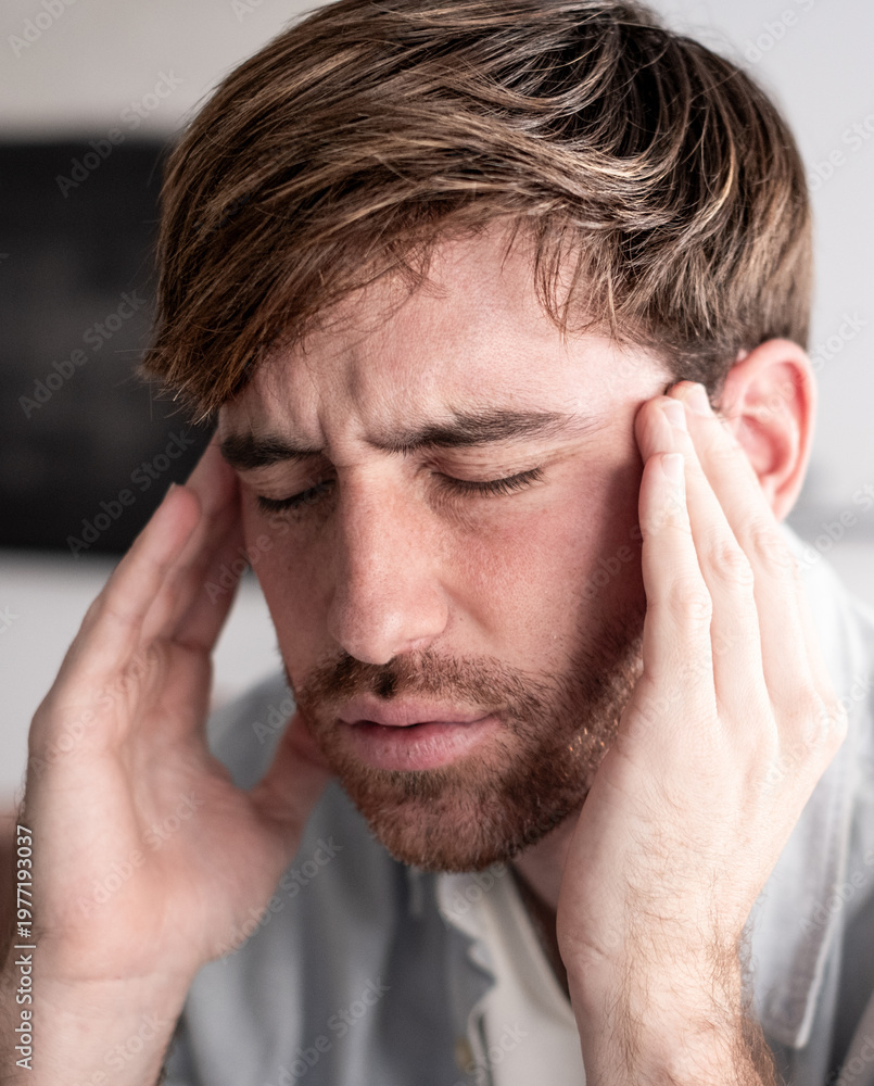 Fototapeta premium Close up portrait of attractive young man with beard sits on the couch at home in a relaxed, modern setting, rubbing his temples to relieve a headache. A bright light comes in from the window.
