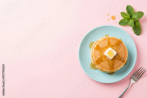 Top view of delicious fluffy pancakes stack with melting butter and sweet maple syrup on a pastel blue plate over a pink background with fresh mint leaves and a silver fork, breakfast food overhead.