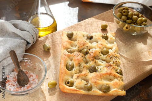 Traditional Italian focaccia with green olives and herbs on wooden cutting board. High-angle shot features salt in glass bowl and olive oil bottle on rustic table. Composition highlights homemade