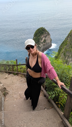 Woman exploring Kelingking Beach in Bali Indonesia, tropical paradise with ocean cliffs and panoramic viewpoint, traveler descending iconic trail, happy raised pose enjoying adventure journey