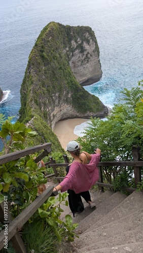 Woman exploring Kelingking Beach in Bali Indonesia, tropical paradise with ocean cliffs and panoramic viewpoint, traveler descending iconic trail, happy raised pose enjoying adventure journey