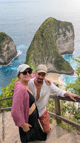 Couple exploring Kelingking Beach in Bali Indonesia, tropical paradise with ocean cliffs and panoramic viewpoint, two people walking down iconic trail enjoying honeymoon adventure, travel journey